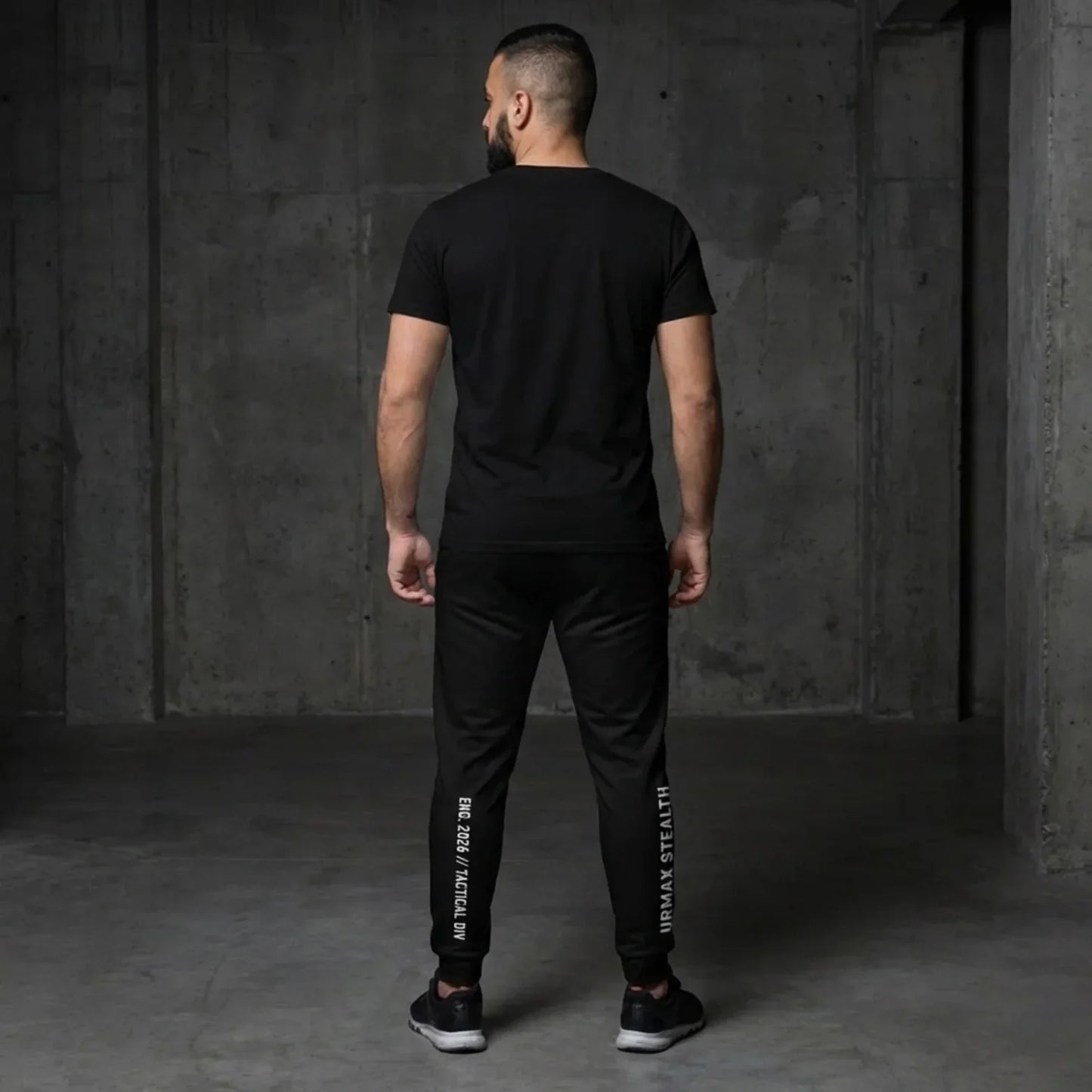 Man in black urmax stealth athletic wear standing in a modern concrete room, rear view.