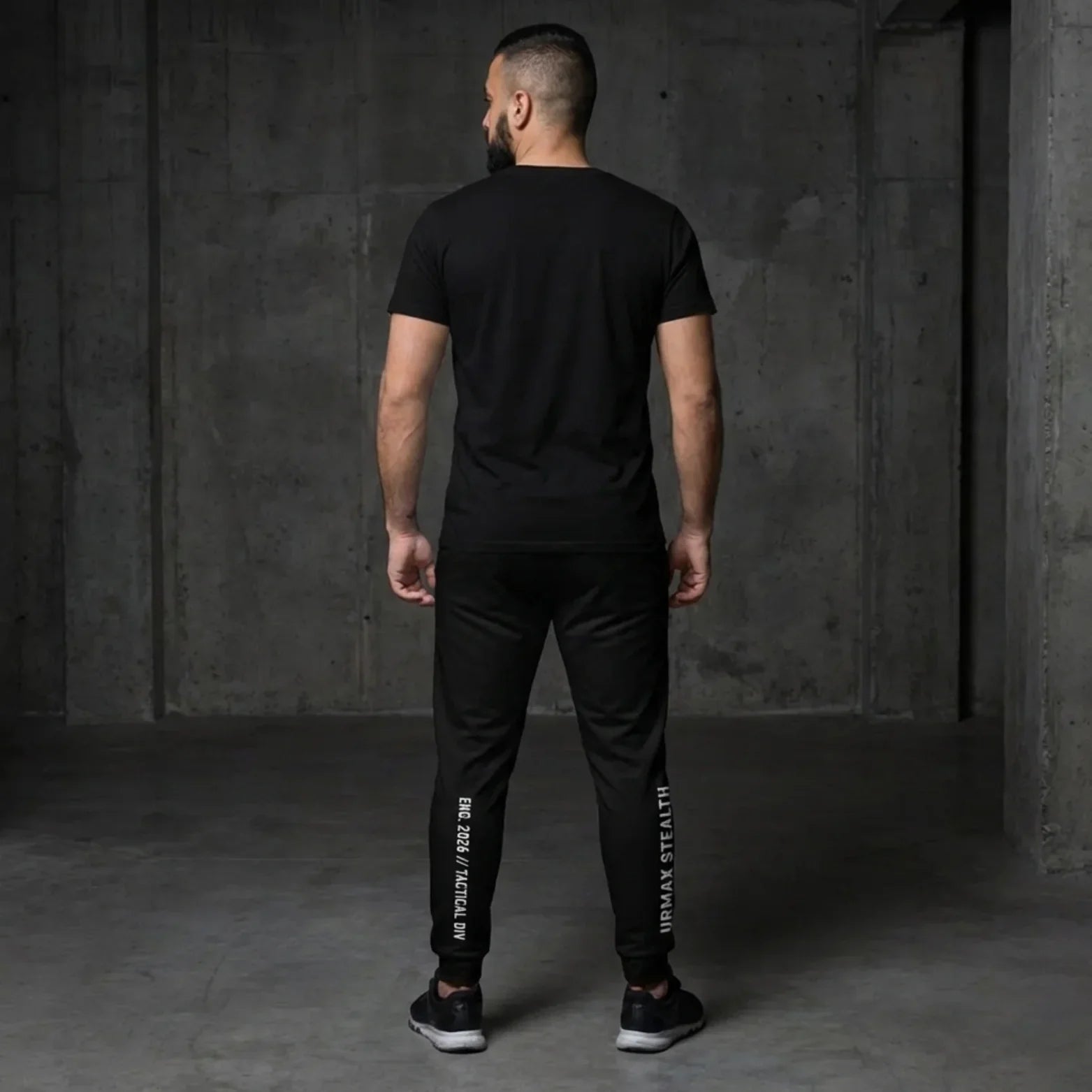 Man in black urmax stealth athletic wear standing in a modern concrete room, rear view.